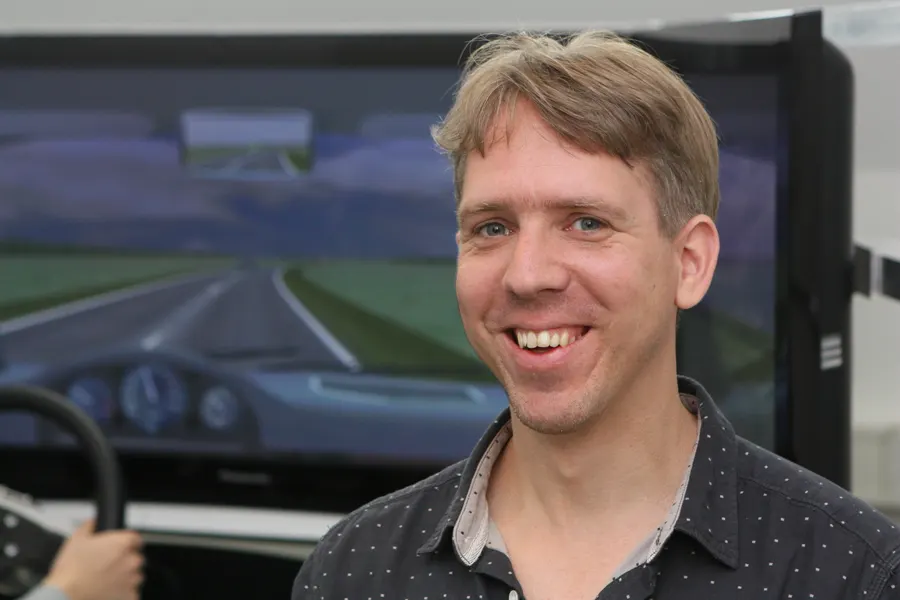 Chris Dijksterhuis lacht in de camera voor het scherm van de rijsimulator